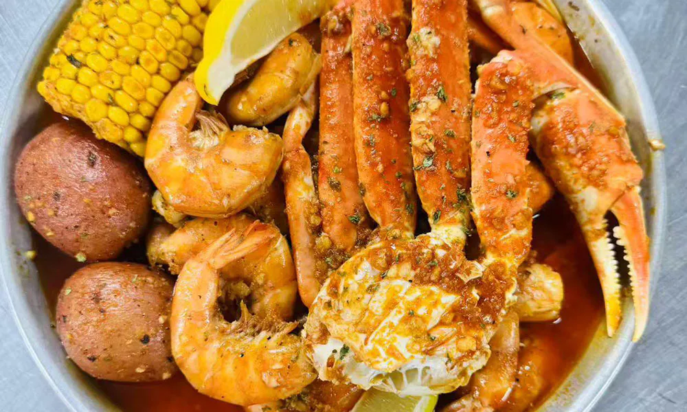 Cajun seafood boil in metal bowl, snow crab legs and shrimp in spicy sauce, served with corn, baby potato and lemon, garnished with garlic herbs