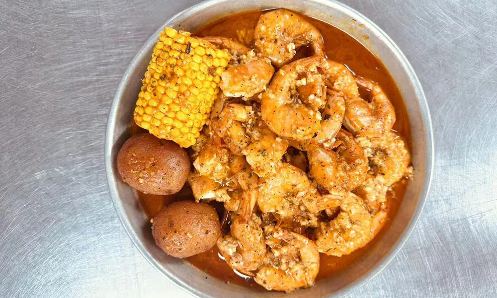 Boiled shrimp in rich spicy Cajun sauce in metal bowl, paired with corn cob and red baby potatoes, seasoned with garlic and herbs on stainless steel surface