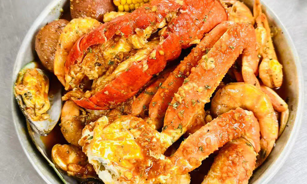 Cajun seafood boil in metal bowl, loaded with lobster tail, crab legs, shrimp and mussels with potato and corn, coated in spicy buttery sauce with herb garnish