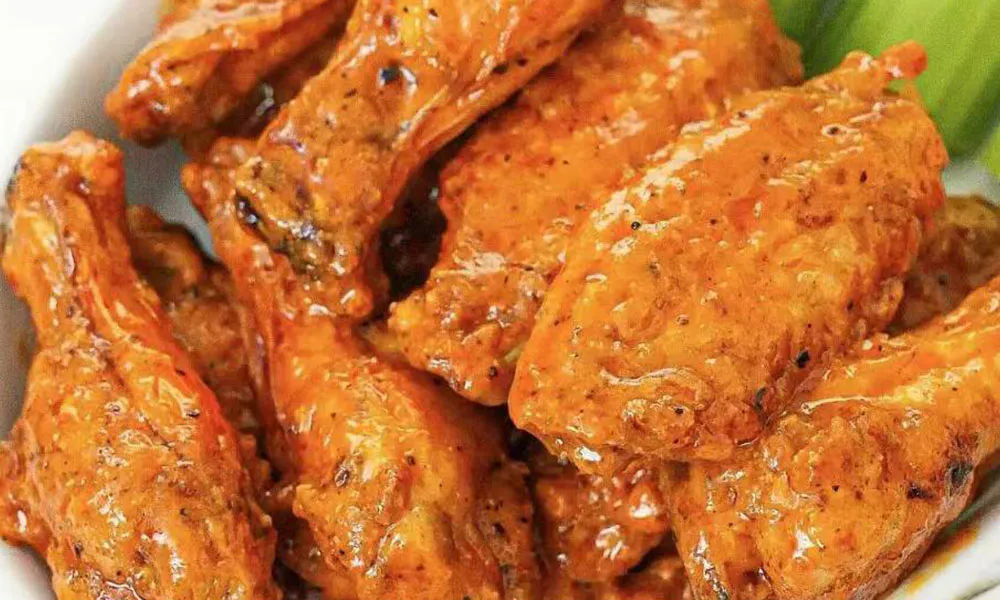 Close-up shot of stacked Buffalo chicken wings covered in glossy orange hot sauce with black pepper, celery sticks partially visible in background