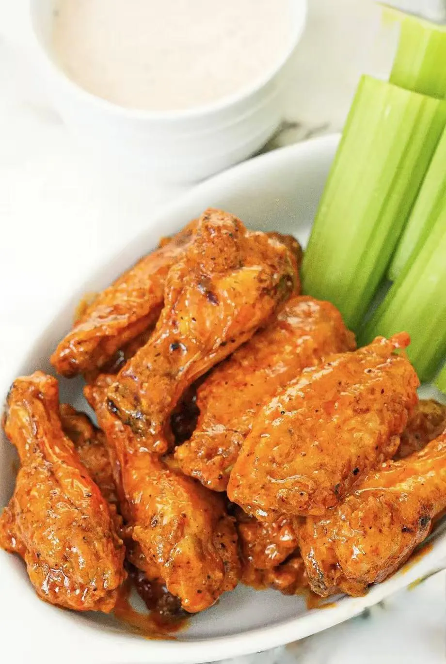 Buffalo chicken wings coated in glossy orange hot sauce on a white oval plate, served with fresh celery sticks and a bowl of creamy dipping sauce on marble countertop